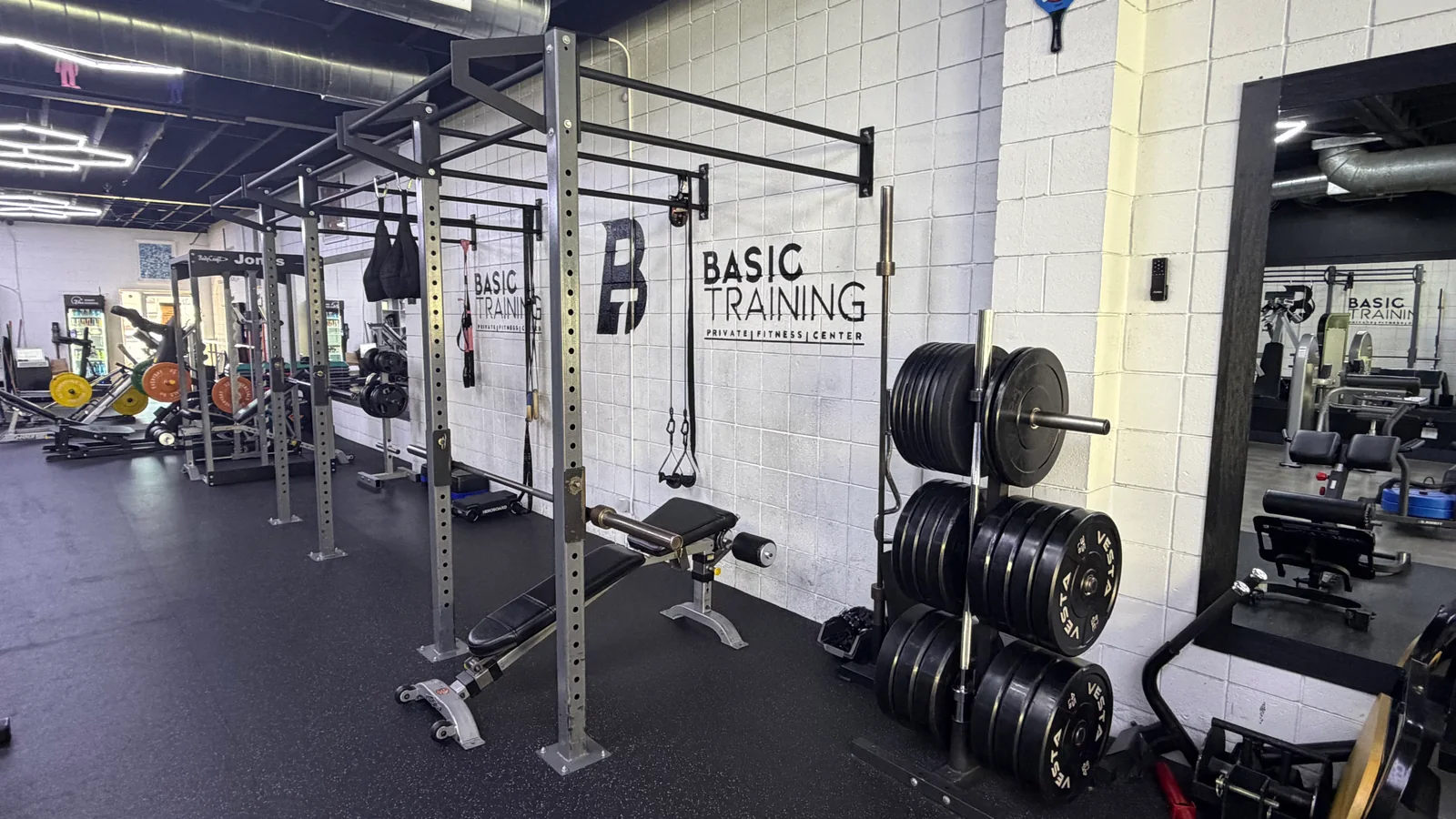 Squat and pull-up rig, barbells and bumper plate storage beside BASIC Training's branded studio wall